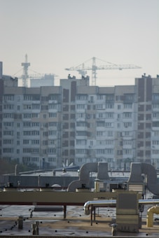 An urban landscape featuring a large residential building with multiple windows and balconies in the background. Several construction cranes rise into the sky, indicating ongoing development or construction. The foreground shows a rooftop with industrial equipment, including vents and ducts.