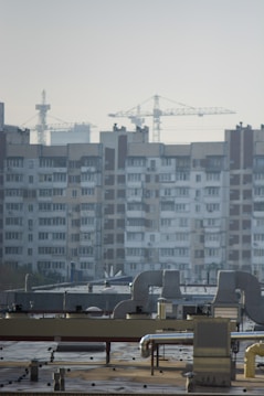 An urban landscape featuring a large residential building with multiple windows and balconies in the background. Several construction cranes rise into the sky, indicating ongoing development or construction. The foreground shows a rooftop with industrial equipment, including vents and ducts.