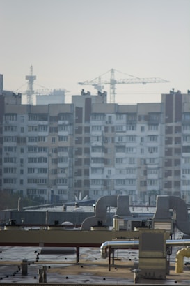 An urban landscape featuring a large residential building with multiple windows and balconies in the background. Several construction cranes rise into the sky, indicating ongoing development or construction. The foreground shows a rooftop with industrial equipment, including vents and ducts.