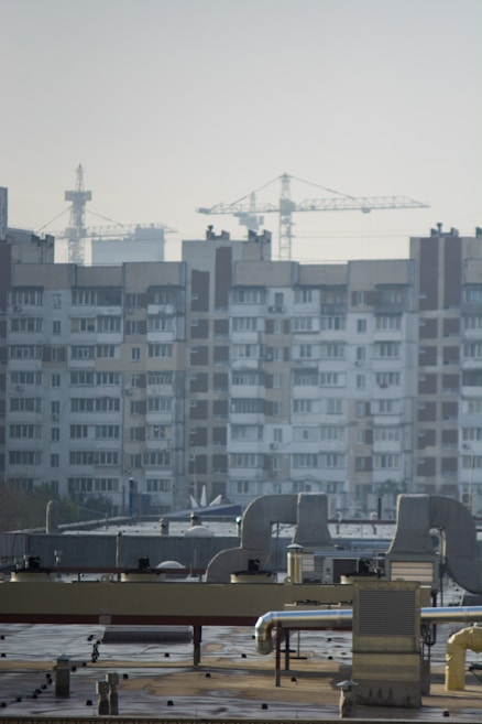 An urban landscape featuring a large residential building with multiple windows and balconies in the background. Several construction cranes rise into the sky, indicating ongoing development or construction. The foreground shows a rooftop with industrial equipment, including vents and ducts.