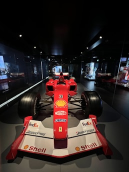 A red Formula 1 racing car is displayed in a dimly lit showroom. The car features prominent branding, including Shell, Magneti Marelli, FedEx, Bridgestone, and others. The racing car has large tires and aerodynamic design, positioned centrally with mirrors reflecting its image on the sides. The dark environment highlights the car's vibrant red color.