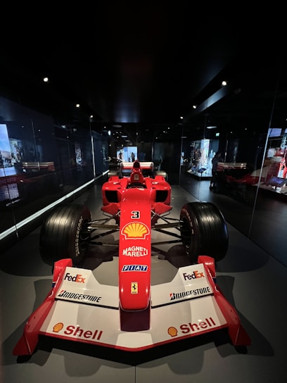A red Formula 1 racing car is displayed in a dimly lit showroom. The car features prominent branding, including Shell, Magneti Marelli, FedEx, Bridgestone, and others. The racing car has large tires and aerodynamic design, positioned centrally with mirrors reflecting its image on the sides. The dark environment highlights the car's vibrant red color.