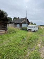 A rustic wooden house with a pitched roof and blue-trimmed windows stands in a grassy rural landscape. A vintage white van is parked beside the house, near a dirt road leading through the greenery. Tall grass and wildflowers surround the buildings, and a wooden fence is visible in the background. The sky is overcast, adding a serene and quaint atmosphere to the scene.