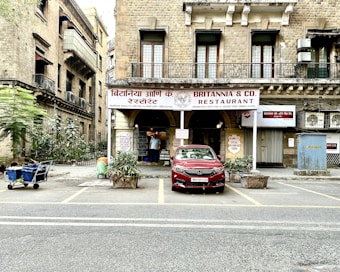 A historical building with a sign reading 'Britannia & Co. Restaurant' in both English and a local language. The facade features large windows and a rustic appearance, with potted plants at the entrance. A red car is parked in front, and a person in traditional attire is visible near the entrance. The street is lined with additional vegetation and a small cart to the side.