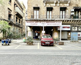 A historical building with a sign reading 'Britannia & Co. Restaurant' in both English and a local language. The facade features large windows and a rustic appearance, with potted plants at the entrance. A red car is parked in front, and a person in traditional attire is visible near the entrance. The street is lined with additional vegetation and a small cart to the side.