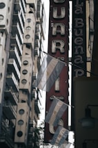 A collection of Argentine flags hanging on a street with tall buildings in the background. The scene includes a vertical red sign with white letters in a vintage font. The lighting suggests an overcast day, creating a subdued atmosphere.