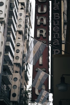 A collection of Argentine flags hanging on a street with tall buildings in the background. The scene includes a vertical red sign with white letters in a vintage font. The lighting suggests an overcast day, creating a subdued atmosphere.