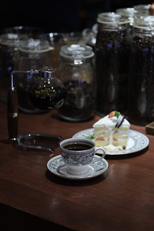 A coffee setup on a wooden table with a vintage-style cup filled with black coffee. A slice of layered cake topped with strawberries and cream is on a decorative plate. In the background, several large glass jars contain coffee beans, adding a rustic elegance to the scene.