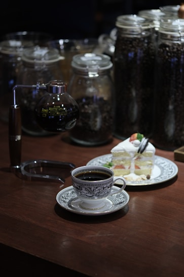 A coffee setup on a wooden table with a vintage-style cup filled with black coffee. A slice of layered cake topped with strawberries and cream is on a decorative plate. In the background, several large glass jars contain coffee beans, adding a rustic elegance to the scene.