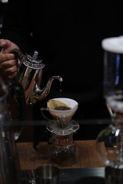 A person is pouring water from a silver gooseneck kettle into a coffee dripper with ground coffee on a wooden surface. The setup includes a glass coffee pot collecting the brewed coffee.