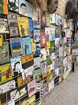 A vibrant display of various artistic tiles featuring colorful illustrations and text in different languages, predominantly Arabic and French. The tiles depict a range of subjects including people, animals, vehicles, and abstract designs. They are arranged in a grid-like pattern on an exterior wall, creating a visually striking collage. Some decorative objects, such as masks, are also visible on the wall.