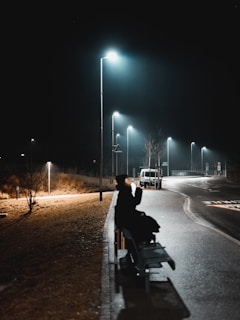 A solitary figure sitting on a park bench under a streetlamp, lost in thought with a cigarette glowing softly in the dark.