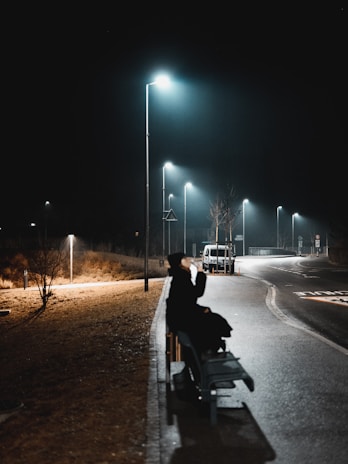 A solitary figure sitting on a park bench under a streetlamp, lost in thought with a cigarette glowing softly in the dark.