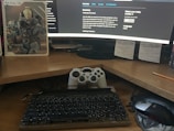 A workspace featuring a curved monitor displaying a software development environment. In front of the monitor is a classic-style mechanical keyboard and a game controller resting against the screen, with a mouse to the side. A notebook with a sci-fi or gaming theme is placed next to the monitor. Sticky notes with handwritten reminders are posted on the wall behind the desk.