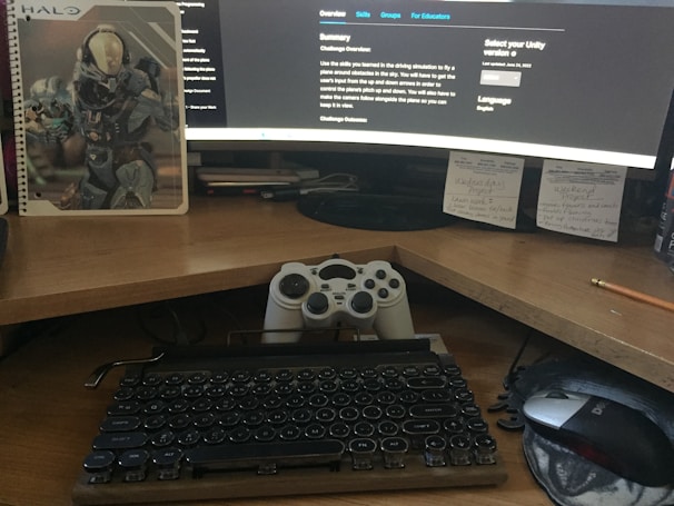 A workspace featuring a curved monitor displaying a software development environment. In front of the monitor is a classic-style mechanical keyboard and a game controller resting against the screen, with a mouse to the side. A notebook with a sci-fi or gaming theme is placed next to the monitor. Sticky notes with handwritten reminders are posted on the wall behind the desk.