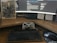A workspace featuring a curved monitor displaying a software development environment. In front of the monitor is a classic-style mechanical keyboard and a game controller resting against the screen, with a mouse to the side. A notebook with a sci-fi or gaming theme is placed next to the monitor. Sticky notes with handwritten reminders are posted on the wall behind the desk.