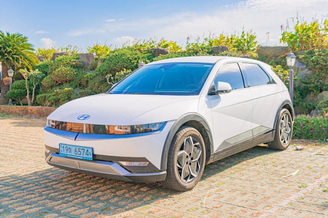 A sleek white electric car is parked on a paved area surrounded by lush greenery and stone-like structures. The modern design features a minimalist aesthetic with sharp lines and a futuristic look. The sunny weather casts clear shadows, enhancing the bright and vibrant scene.