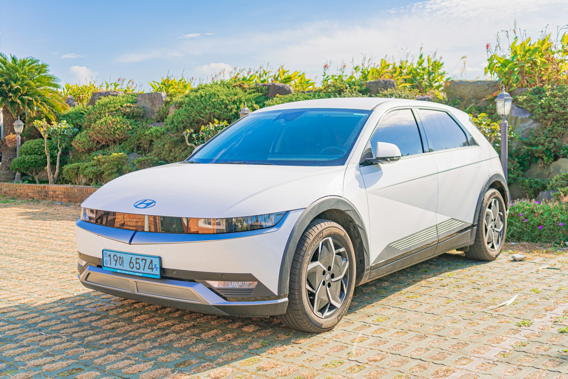 A sleek white electric car is parked on a paved area surrounded by lush greenery and stone-like structures. The modern design features a minimalist aesthetic with sharp lines and a futuristic look. The sunny weather casts clear shadows, enhancing the bright and vibrant scene.