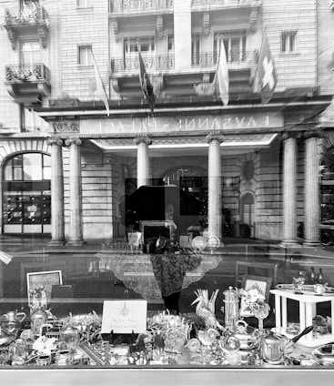 A shop window displays various decorative items, including silverware and glassware, with reflections of a grand building featuring columns and flags in the background. The window reveals an elegant and luxurious atmosphere.