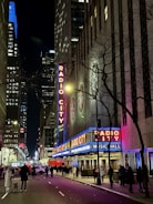 A nighttime street scene of a bustling city with neon lights illuminating a building labeled 'Radio City Music Hall.' The street is lined with pedestrians and vehicles, framed by tall skyscrapers. The lights create a vibrant and lively atmosphere.