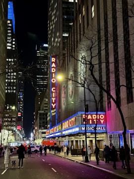 A nighttime street scene of a bustling city with neon lights illuminating a building labeled 'Radio City Music Hall.' The street is lined with pedestrians and vehicles, framed by tall skyscrapers. The lights create a vibrant and lively atmosphere.