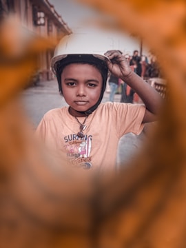 A young child is wearing a helmet and a t-shirt with a surfing design. The background is a street setting, with other people possibly present in a blurred focus. The foreground features an artistic frame with warm tones, creating an interesting visual effect.