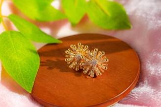 Gold flower-shaped earrings rest on a round wooden platform, surrounded by lush green leaves and a soft pink fabric background.