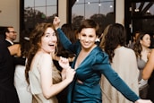 Two women are smiling and dancing at a party or social gathering. They are surrounded by other people who are also dressed in formal or semi-formal attire. The atmosphere appears lively and joyful, with large windows and lights visible in the background.