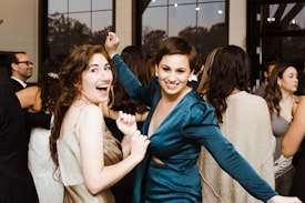 Two women are smiling and dancing at a party or social gathering. They are surrounded by other people who are also dressed in formal or semi-formal attire. The atmosphere appears lively and joyful, with large windows and lights visible in the background.