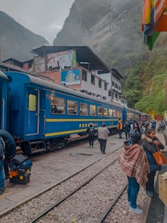 A blue train is stationed on a track with several people gathered nearby, some carrying backpacks and wearing jackets. The setting appears to be in a mountainous area, with mist partially obscuring the surrounding landscape. Buildings with signs are visible in the background.