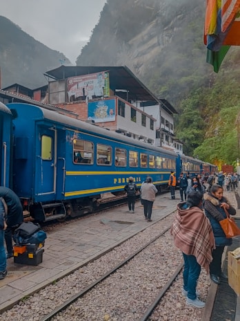 A blue train is stationed on a track with several people gathered nearby, some carrying backpacks and wearing jackets. The setting appears to be in a mountainous area, with mist partially obscuring the surrounding landscape. Buildings with signs are visible in the background.
