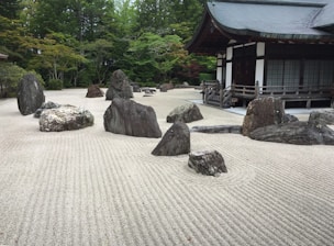 A traditional Japanese Zen garden featuring carefully arranged rocks of various sizes on meticulously raked gravel. The rocks are positioned to create a harmonious and serene atmosphere. In the background, a traditional Japanese building with wooden architecture and a tiled roof adds to the peaceful setting. Surrounding the garden are lush green trees, contributing to the tranquil environment.