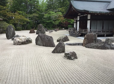 A traditional Japanese Zen garden featuring carefully arranged rocks of various sizes on meticulously raked gravel. The rocks are positioned to create a harmonious and serene atmosphere. In the background, a traditional Japanese building with wooden architecture and a tiled roof adds to the peaceful setting. Surrounding the garden are lush green trees, contributing to the tranquil environment.