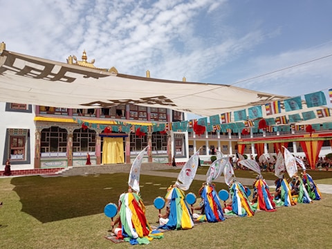 A group of people in colorful ceremonial attire are performing a traditional dance with large ornate fans. They are situated in a courtyard of a building with intricate architectural details and vibrant decorations. Strings of multi-colored flags hang above, and the sky is clear with scattered clouds.