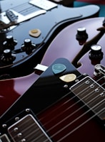 Two electric guitars are lying close together, with a focus on the polished, glossy surfaces. Various control knobs and strings are visible, and a couple of guitar picks rest on top of one guitar.