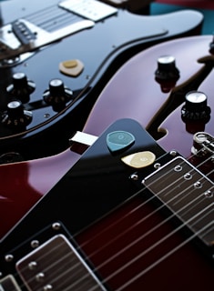 Two electric guitars are lying close together, with a focus on the polished, glossy surfaces. Various control knobs and strings are visible, and a couple of guitar picks rest on top of one guitar.