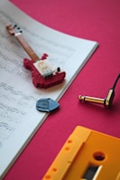An open sheet of music lies on a red surface with a small toy guitar made of interlocking bricks placed on top. Next to the sheet music is a black guitar pick and an audio jack connector. A yellow cassette tape is partially visible on the red background.