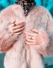 A close-up of someone wearing a luxurious, fluffy pink fur coat and multiple rings on their fingers. A decorative scarf with a floral pattern adds contrast to the soft texture of the coat.