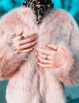 A close-up of someone wearing a luxurious, fluffy pink fur coat and multiple rings on their fingers. A decorative scarf with a floral pattern adds contrast to the soft texture of the coat.