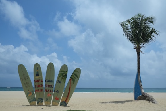 Multiple surfboards with colorful designs are planted upright in the sand, forming a decorative welcome sign on a pristine beach. A solitary palm tree stands next to the surfboards against a backdrop of calm blue sea and a partly cloudy sky.