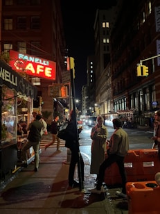 A nighttime street scene in a city with people gathered outside a cafe. Bright neon signage illuminates the scene, contrasting with the dark surroundings. Several people are engaged in conversation, some holding drinks. Orange traffic barriers are visible on the right, alongside parked cars lining the dimly lit street.