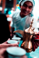 A person with a towel or headscarf smiles while serving traditional Arabic coffee using a brass dallah, a traditional coffee pot. Two small cups are held in the foreground, ready to be filled.