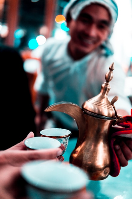 A person with a towel or headscarf smiles while serving traditional Arabic coffee using a brass dallah, a traditional coffee pot. Two small cups are held in the foreground, ready to be filled.