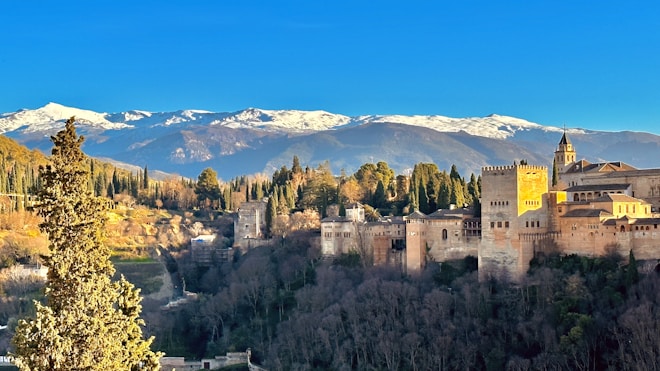 A historic fortress with intricate architecture is set against a backdrop of lush green trees and vast mountains with snow-covered peaks. The clear blue sky enhances the serene and majestic atmosphere of the scene.