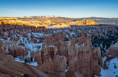 A stunning landscape featuring towering rock formations known as hoodoos in Bryce Canyon National Park. The formations are covered with patches of snow, contrasting with the warm orange and red hues of the rock. Sunlight casts a golden glow on some of the formations, highlighting their unique shapes. A vast view of distant plateaus and ridges can be seen in the background under a clear blue sky.