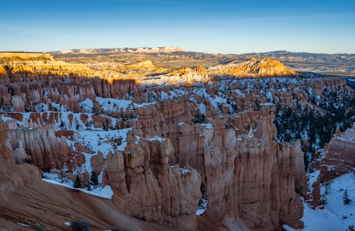A stunning landscape featuring towering rock formations known as hoodoos in Bryce Canyon National Park. The formations are covered with patches of snow, contrasting with the warm orange and red hues of the rock. Sunlight casts a golden glow on some of the formations, highlighting their unique shapes. A vast view of distant plateaus and ridges can be seen in the background under a clear blue sky.