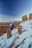 Snow-covered red sandstone hoodoos rise majestically in a steep canyon under a clear blue sky. A narrow trail winds through the landscape, bordered by tall, rugged formations with patches of snow clinging to their surfaces. The sunlight casts warm hues on the rocky structures, highlighting their intricate details.