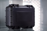 A sturdy black protective case with a rectangular shape and reinforced edges, set against a reflective surface. The case features multiple latches and a secure handle for easy transport, with a simple industrial design.