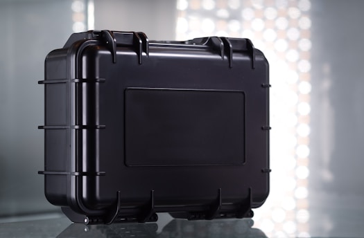 A sturdy black protective case with a rectangular shape and reinforced edges, set against a reflective surface. The case features multiple latches and a secure handle for easy transport, with a simple industrial design.
