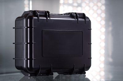 A sturdy black protective case with a rectangular shape and reinforced edges, set against a reflective surface. The case features multiple latches and a secure handle for easy transport, with a simple industrial design.